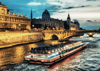 An illuminated river cruise ship gliding through a city’s waterways with historic architecture in the background at dusk.