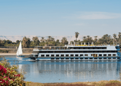 A luxury cruise ship sailing along the calm Nile River, surrounded by desert scenery and vibrant flowers.