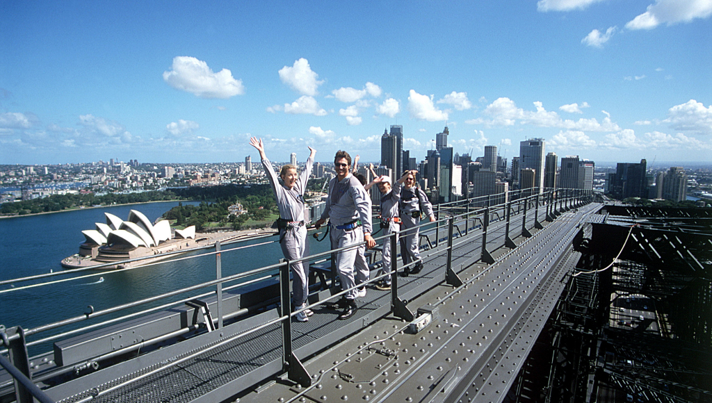 Travelers climbing the Sydney Harbour Bridge with the Sydney Opera House in the background on a sunny day