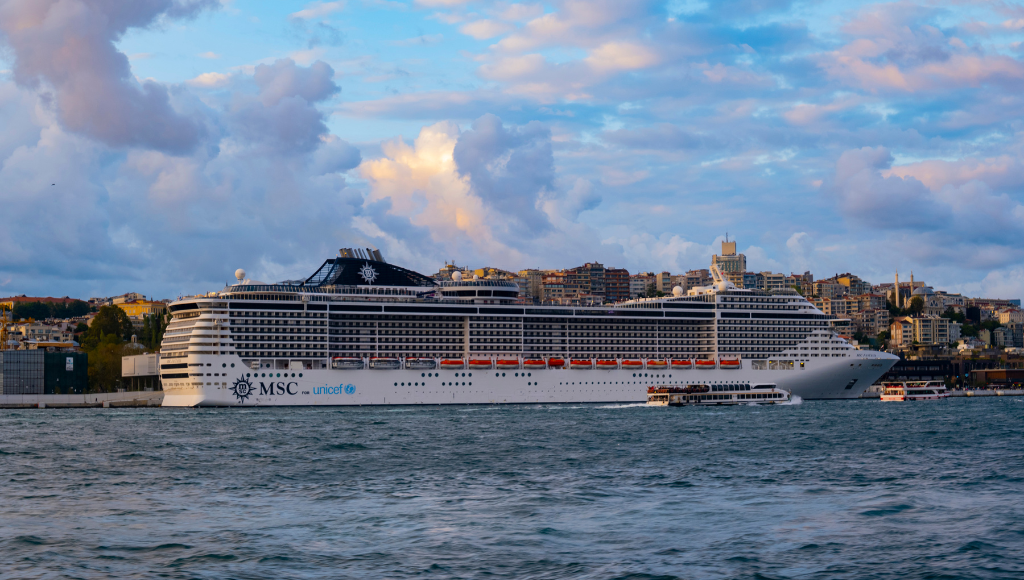 An MSC cruise ship docked at a port with a city skyline and cloudy sky in the background.