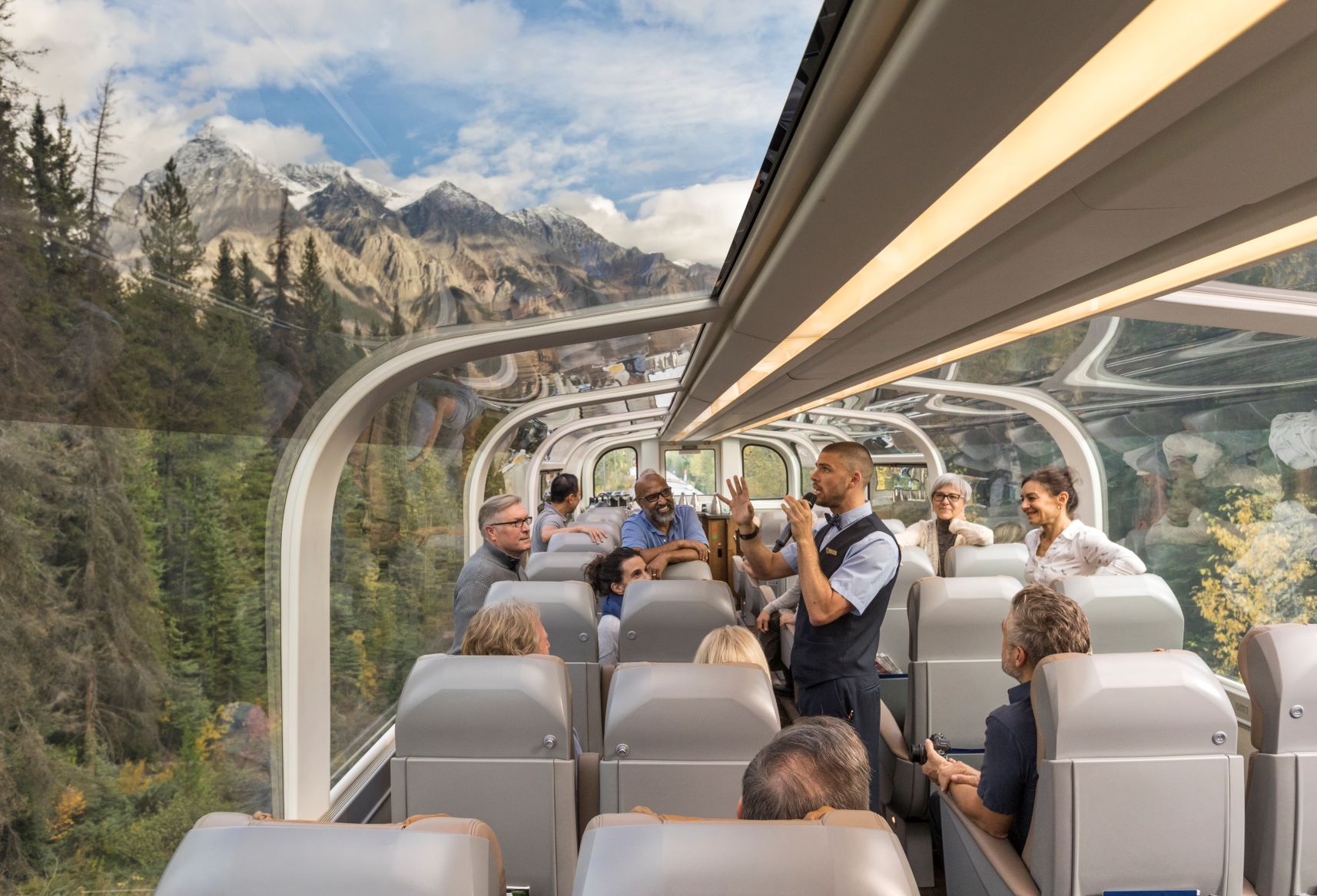 Passengers enjoying a luxury rail journey in a glass-domed train car, surrounded by stunning mountain and forest views, with a guide sharing insights.