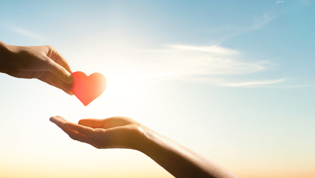 One hand passing a red heart to another hand against a sunset sky, symbolizing the act of giving and charity.